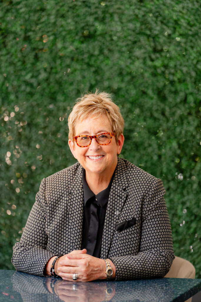 Catherine Gray Sitting with hands crossed in front of green foliage background