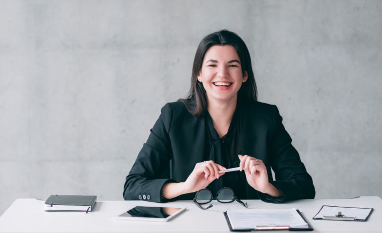 Woman at desk in black suit smiling and holding pen