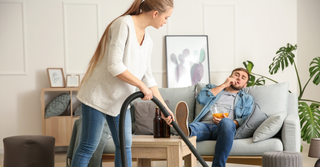 woman vacuuming with man sitting on couch eating chips