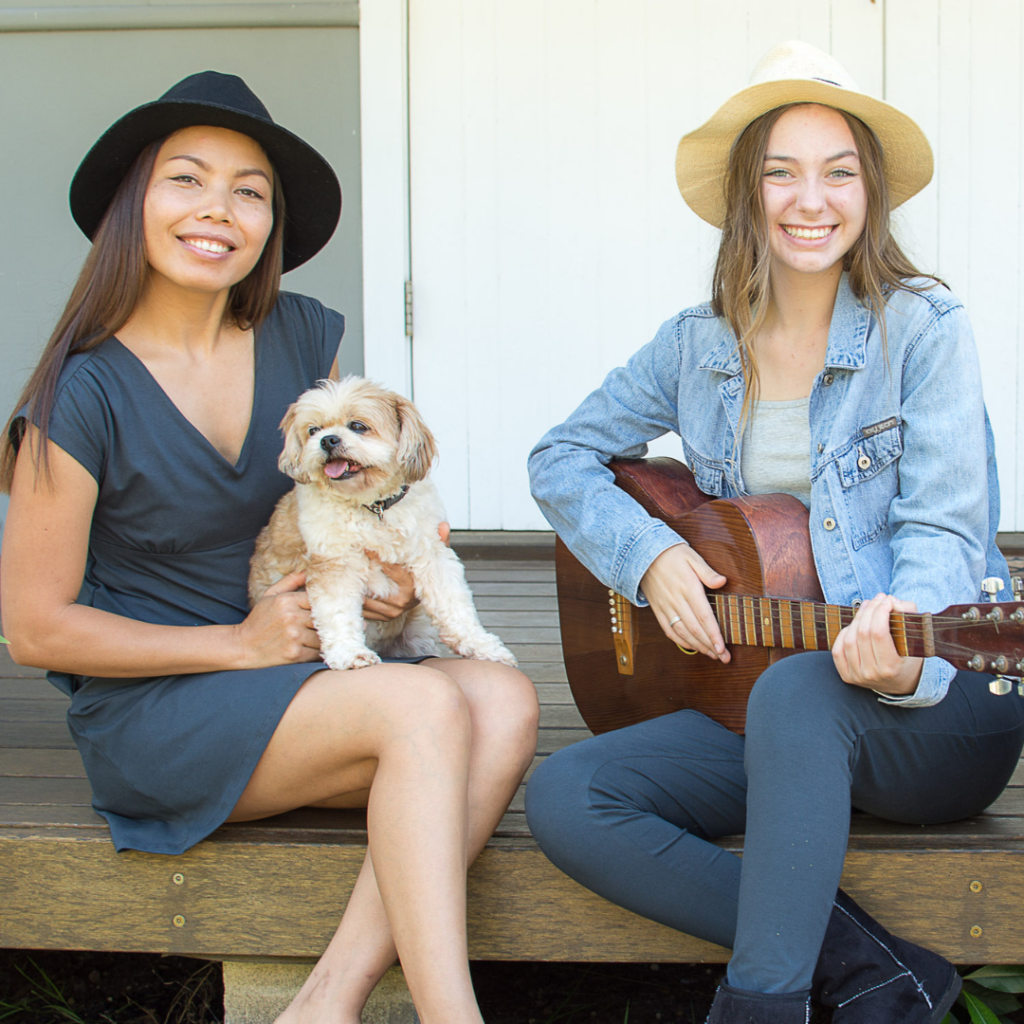 two girls sitting on deck, one holding dog and one holding guitar